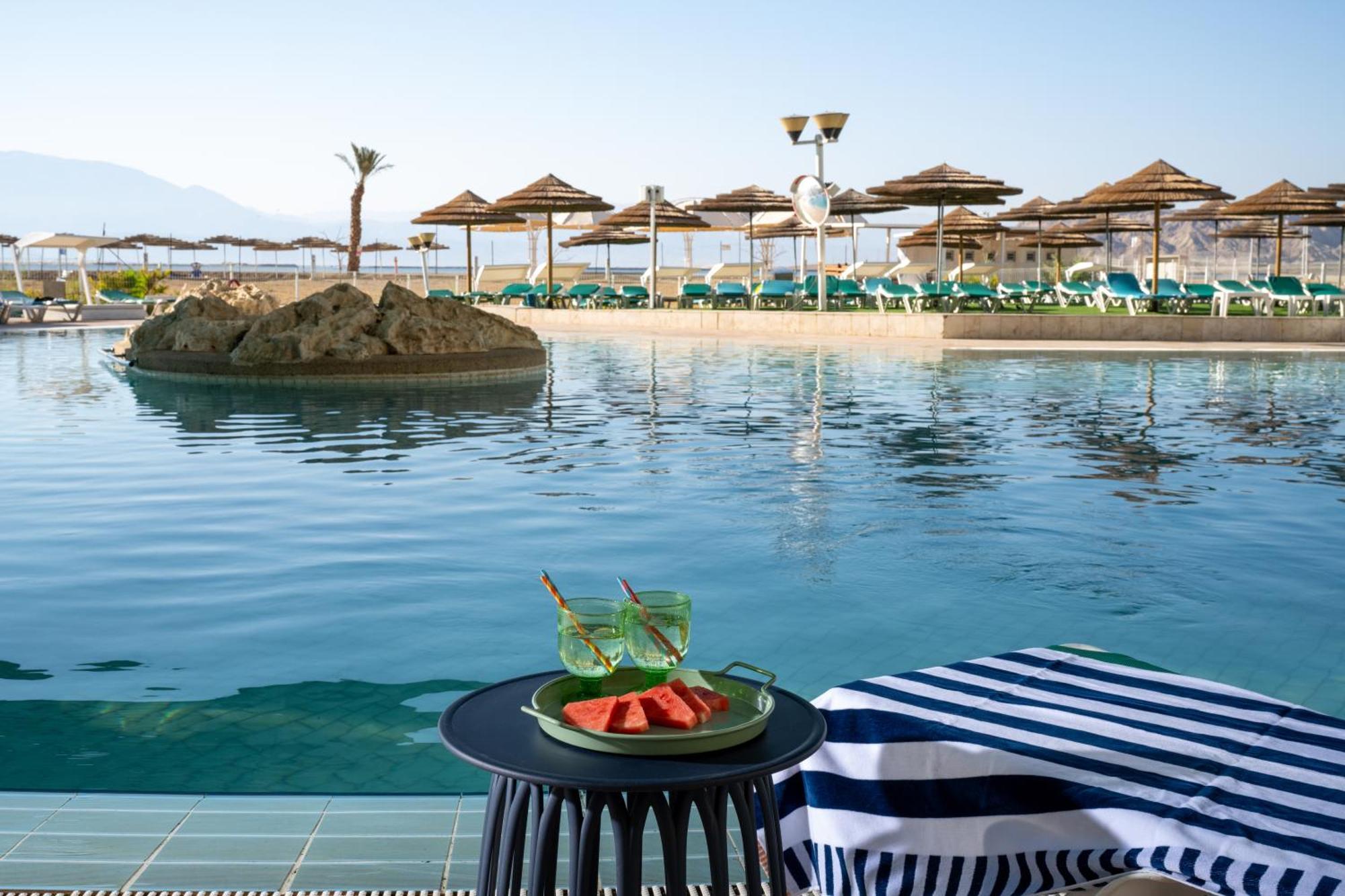 Leonardo Club Hotel Dead Sea - All Inclusive Neve Zohar Exterior foto The photo depicts a tranquil swimming pool area at a resort or hotel. In the foreground, there is a small round table with a plate containing slices of watermelon and two green drinks. The background features a calm pool with a small rock island in t