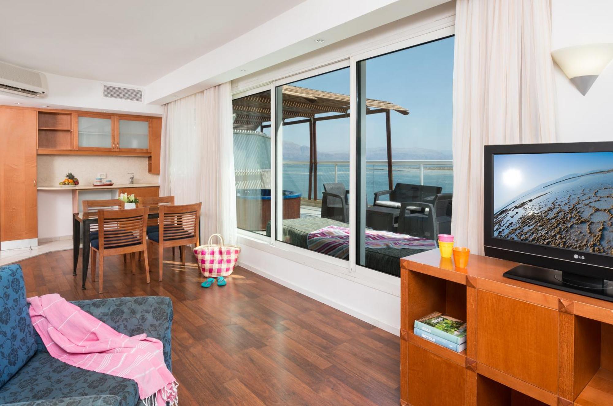Leonardo Club Hotel Dead Sea - All Inclusive Neve Zohar Exterior foto The photo depicts a modern living space with a view. On one side, there is a comfortable seating area with a couch and a television cabinet. The TV is turned on, showing a scenic image. 

In the background, there is a dining area with a wooden table 